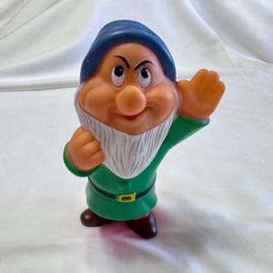 May include: A molded rubber figurine of a character with a blue hat, peach-colored skin, and a white beard. The character wears a green tunic with a black belt featuring a yellow buckle and brown shoes. One hand is raised in a wave gesture.