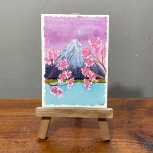 Op de afbeelding: Een rechthoekig aquarel op een kleine houten ezel toont een berg met een besneeuwde top onder een paarse lucht met een maansikkel. Roze kersenbloesemtakken omlijsten de berg boven een lichtblauw wateroppervlak.