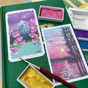 Peut inclure: Deux aquarelles sur papier blanc. La première peinture représente une montagne avec des fleurs de cerisier roses au premier plan. La deuxième peinture représente un coucher de soleil sur un lac avec deux arbres au premier plan. Les deux peintures sont sur un livre vert avec des godets d'aquarelle et un pinceau.