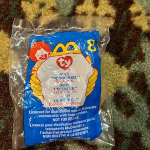 May include: A blue and white Ty Beanie Baby packaging with a picture of Ronald McDonald holding a Ty Beanie Baby. The packaging is for the "Nuts the Squirrel" Beanie Baby. The packaging says "Licensed for distribution only by McDonald's restaurants with food purchase. Not for resale."