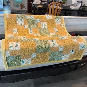 Puede incluir: Un edredón hecho a mano con un diseño de patchwork. El edredón presenta cuadrados de tela amarilla, verde y blanca. Algunos cuadrados tienen un patrón de ciervos. El edredón se exhibe en un marco de acolchado, mostrando su tamaño y artesanía.