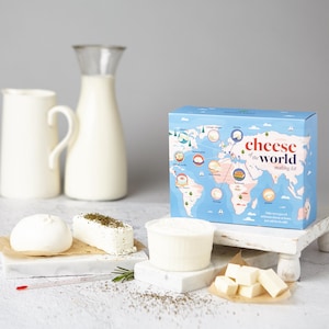 May include: A blue Cheese of the World making kit box featuring a world map, accompanied by a white pitcher, a round cheese, a rectangular herb-topped cheese, a tub of white cheese, and several small white cheese cubes on a white surface.