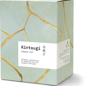 May include: A rectangular box featuring a Kintsugi repair kit. The box has a light green background with gold-colored crack patterns. The text "Kintsugi repair kit" is displayed on a white label, along with Japanese characters and the phrase "Celebrate imperfection by repairing your own ceramics with gold."