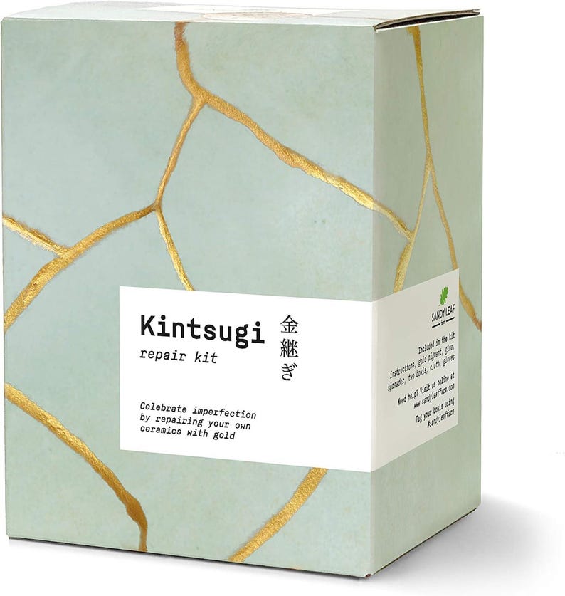 May include: A rectangular box featuring a Kintsugi repair kit. The box has a light green background with gold-colored crack patterns. The text "Kintsugi repair kit" is displayed on a white label, along with Japanese characters and the phrase "Celebrate imperfection by repairing your own ceramics with gold."