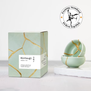 May include: A Kintsugi repair kit box in light green with gold crack patterns, featuring the text Kintsugi repair kit and Celebrate imperfection by repairing your own ceramics with gold. Beside it, two small light green bowls with gold repair lines.