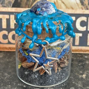 May include: A small glass jar with a blue wax seal and a star charm. The jar is filled with black sand, dried flowers, and other natural elements.