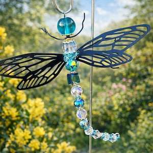May include: A decorative dragonfly garden stake. The dragonfly features black metal wings with intricate cut-out designs. The body is adorned with blue and clear beads, and a teal glass orb. The stake is made of silver metal.