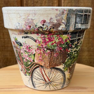 May include: Decorative flower pot with a painted design featuring a red bicycle with a basket full of pink flowers. The pot has a distressed, aged appearance, with a cream-colored rim and a scene of a building with a window.