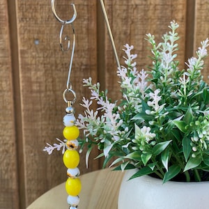 May include: A decorative metal plant hanger with a beaded charm. The charm features yellow and white beads, with a sunflower design on one bead. The hanger is silver and is designed to hold a small plant pot.