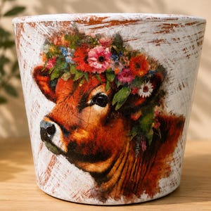 May include: A tapered cylindrical plant pot with a white distressed finish featuring a printed illustration of a brown cow wearing a vibrant floral crown made of red, pink, orange, and blue flowers with green leaves on the front surface.