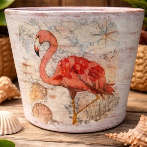 May include: A decorative, off-white ceramic planter featuring a flamingo illustration. The planter has a distressed finish with a map design and seashell accents. The flamingo is depicted in shades of pink and red.