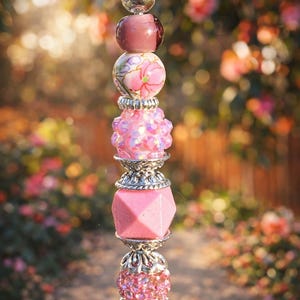 May include: A decorative beaded charm with a silver chain and various pink beads. The beads include a floral-painted bead, a faceted pink bead, and a bead with pink crystals. The charm is suspended against a blurred background of flowers.