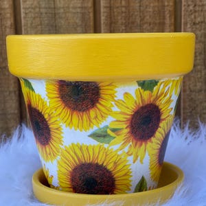 May include: A yellow painted flower pot with a matching saucer. The pot is decorated with a pattern of sunflowers in full bloom. The rim and saucer are a solid yellow, creating a bright and cheerful look.