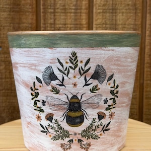 May include: A decorative flower pot with a distressed, whitewashed finish. The pot features a detailed illustration of a bumblebee surrounded by floral and leaf elements. The rim is painted green and gold. The pot is designed for planting.