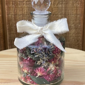 May include: A clear glass bottle filled with dried flowers and herbs, topped with a faceted crystal stopper. A cream-colored ribbon is tied around the neck of the bottle. The flowers are shades of pink, purple, and green.