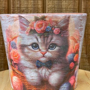 May include: A decorative ceramic pot featuring a detailed illustration of a fluffy kitten with blue eyes, wearing a floral crown and a blue bow tie. The kitten is nestled in a woven basket with yarn and flowers, perfect for a plant or as a decorative piece.