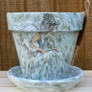 May include: A white ceramic planter with a painted image of a fairy with wings and a flower crown. The planter has a matching saucer.