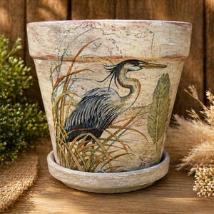 May include: A decorative beige ceramic flower pot with a matching saucer. The pot features a detailed illustration of a blue heron standing in tall grass. The design has a vintage, weathered look, with a natural, earthy aesthetic.