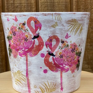 May include: White ceramic planter with a distressed finish, featuring two pink flamingos with floral bouquets. Gold palm leaves and butterflies accent the design. A decorative container for plants.
