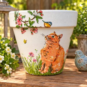 May include: A white ceramic plant pot featuring a hand-painted orange tabby cat sitting in green grass with pink flowers. Above the cat, a small blue and yellow bird perches on a branch with pink blossoms, and a yellow butterfly flies nearby.