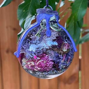 May include: A purple glass ornament filled with dried flowers and a silver key charm. The ornament is hanging from a purple string.