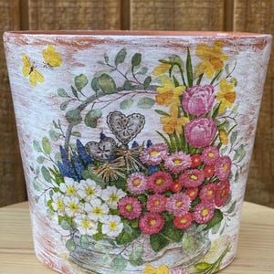May include: A terracotta-colored flower pot with a distressed white finish. The pot is decorated with a floral design featuring pink, yellow, and white flowers, green leaves, and a yellow butterfly. The pot is sitting on a wooden surface.