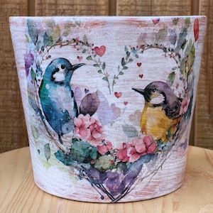 May include: A decorative ceramic planter with a heart-shaped floral design featuring two birds. The planter has a distressed white background with pink, purple, and green accents. The birds are blue and yellow.