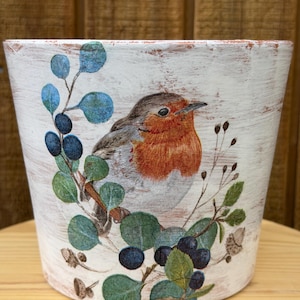 May include: White distressed ceramic flower pot with a painted design of a robin bird, surrounded by green leaves, blue berries, and sprigs. The pot has a rustic, farmhouse style and is suitable for indoor or outdoor use.