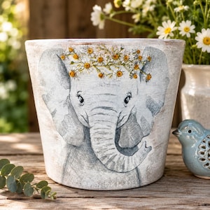 May include: A tapered ceramic planter featuring a hand-painted illustration of a grey elephant calf wearing a crown of yellow and white daisies. The pot has a distressed white finish and a wide rim, showcasing a detailed animal portrait.