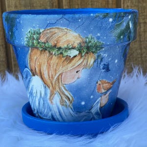 May include: Blue ceramic flower pot with a matching saucer. The pot features a winter scene with an angel, a bird, and a wreath. The angel has blonde hair and wears a white gown. The pot is approximately 5 inches tall.