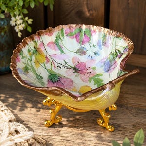 May include: A decorative bowl shaped like a seashell, featuring a white interior with a colorful floral pattern in shades of pink, yellow, and blue. The rim is finished in a metallic bronze tone, and the bowl rests on a gold-colored metal stand.