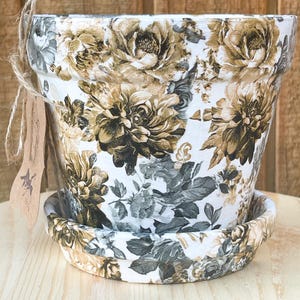 May include: A white ceramic planter with a floral pattern in shades of brown, gray, and beige. The planter has a matching saucer.