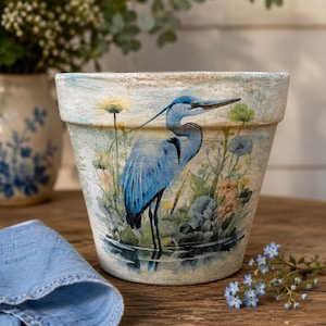 May include: A tapered terracotta plant pot featuring a painted illustration of a blue heron standing in water surrounded by aquatic plants and flowers. The pot has a textured, weathered finish with light blue and cream tones throughout the surface.