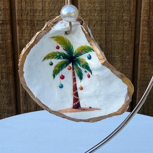 May include: A decorative oyster shell ornament featuring a hand-painted palm tree adorned with colorful Christmas ornaments and string lights. The shell has a gold-painted edge and is suspended by a jute twine string with a single white pearl bead.