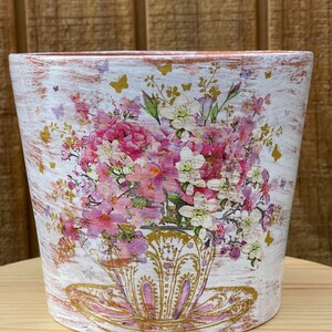 May include: A white, distressed-finish flower pot with a floral design. The pot features a bouquet of pink and white flowers in a decorative teacup, with gold accents and butterfly details. The pot is cylindrical and sits on a wooden surface.