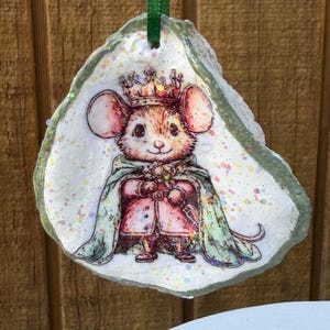 May include: An oyster shell ornament featuring a printed illustration of a mouse wearing a crown and a green cape. The shell has a white base with iridescent glitter accents and is suspended by a green ribbon loop for hanging purposes.