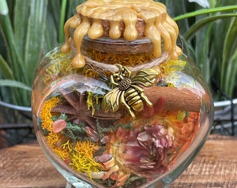 Può includere: Un barattolo di vetro a forma di cuore riempito di fiori secchi, erbe e un ciondolo a forma di ape dorata. Il barattolo ha un tappo di sughero con un sigillo di cera dorata.