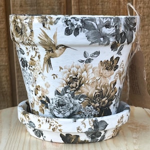 May include: White terracotta planter with a floral pattern in shades of brown, gray, and white. A hummingbird is flying in the center of the planter. The planter has a matching saucer.