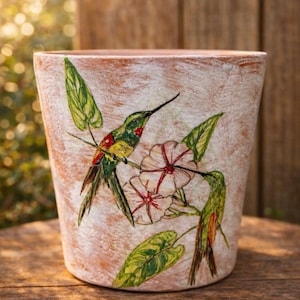 May include: A terracotta-colored ceramic flower pot with a distressed white wash. The pot is decorated with a colorful illustration of hummingbirds, flowers, and green leaves. The pot is sitting on a wooden surface.