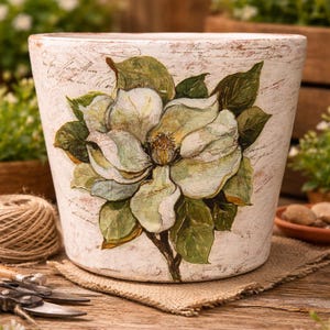 May include: A white ceramic flower pot with a distressed finish, featuring a hand-painted magnolia flower design in shades of green and cream. The pot sits on a burlap mat, suggesting a garden theme.