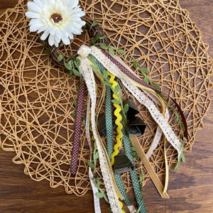 May include: A decorative wreath with a large white daisy, green leaves, and various ribbons. Ribbons include white lace, brown with white polka dots, and gold. The wreath is on a woven placemat and a dark wooden surface.