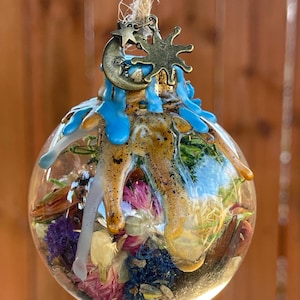 May include: A clear glass orb ornament filled with dried flowers in shades of purple, pink, and yellow. The ornament has a bronze-colored charm with a crescent moon, star, and sun design. Blue resin drips down the top of the orb, which hangs from a natural twine.