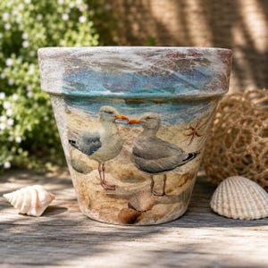 May include: A tapered terracotta plant pot featuring a decorative decoupage print of two seagulls standing on a sandy beach with seashells and a starfish. The upper rim has a distressed white and blue wash finish over the natural clay surface.