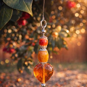 May include: A decorative ornament with a heart-shaped amber-colored bead, suspended from a silver-colored metal hook. The ornament also features a red and orange bead, and a round orange bead. The background is blurred with natural light.