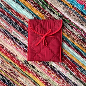 May include: A red quilted fabric pouch with a red ribbon tie. The pouch is lying on a colorful striped rug.