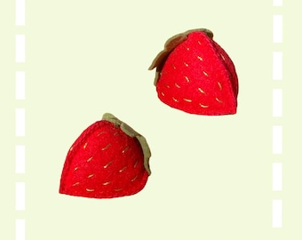 May include: Two red felt strawberries with gold stitching. The strawberries are on a light green background.