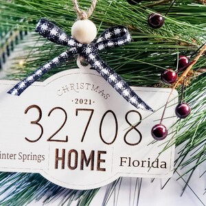 Op de afbeelding: Witte houten kerstversiering met zwart-wit geruit lint, een houten kraal en de tekst "Christmas 2021", "32708", "Winter Springs", "HOME" en "Florida".