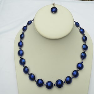Peut inclure: Un collier de perles bleu fonc&eacute; et un pendentif assorti. Le collier comporte de grosses perles rondes entrecoup&eacute;es de perles ovales plus petites. Le pendentif est une seule perle ronde. Les bijoux sont expos&eacute;s sur un buste blanc.