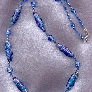 May include: A blue and white glass bead necklace with a floral pattern. The necklace is made of small blue glass beads and larger, oval-shaped beads with a floral design.