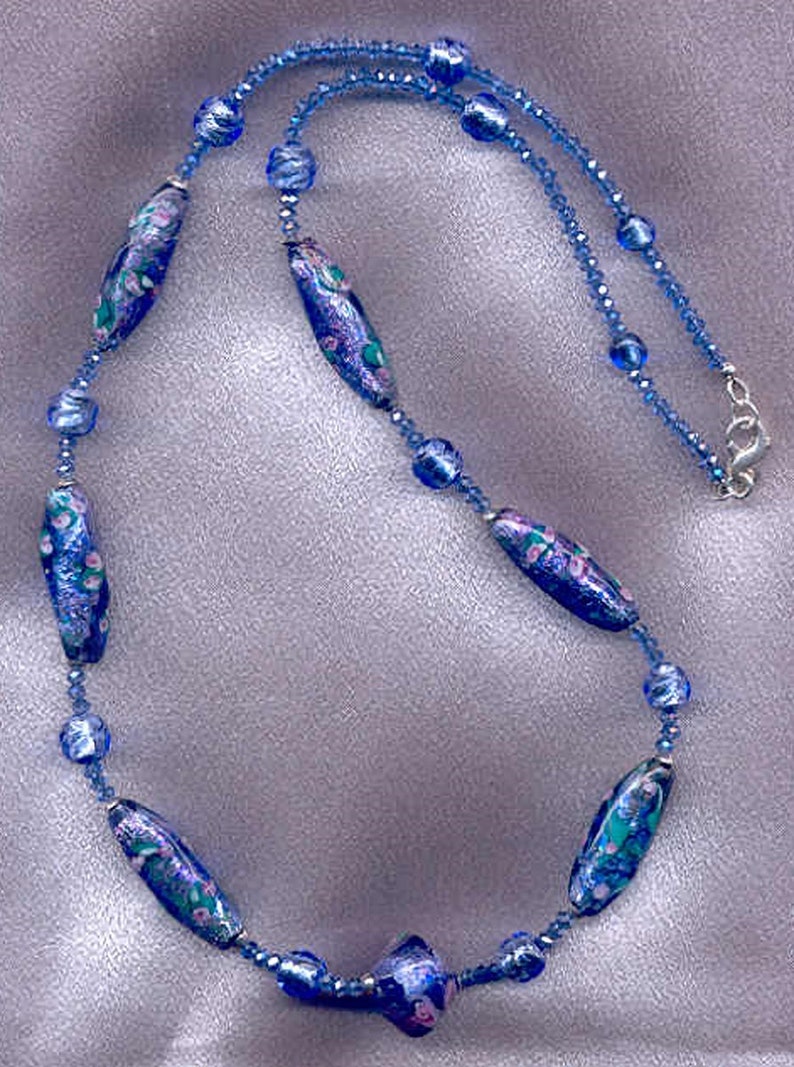 May include: A blue and white glass bead necklace with a floral pattern. The necklace is made of small blue glass beads and larger, oval-shaped beads with a floral design.
